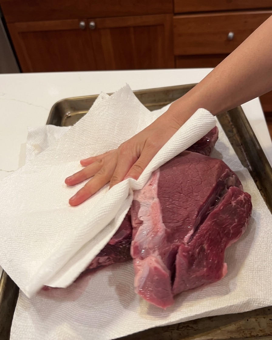 A hand using a white paper towel to pat a large piece of raw beef dry on a baking sheet.