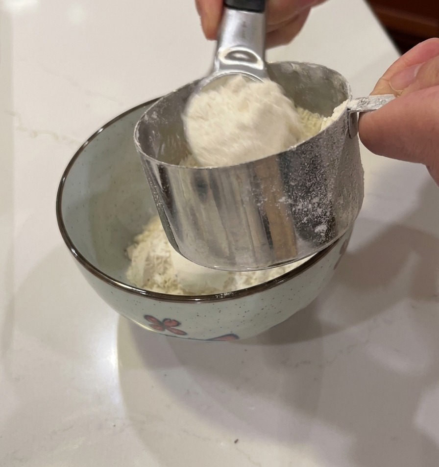 Hands pouring white flour from a metal measuring cup into a small, patterned bowl.