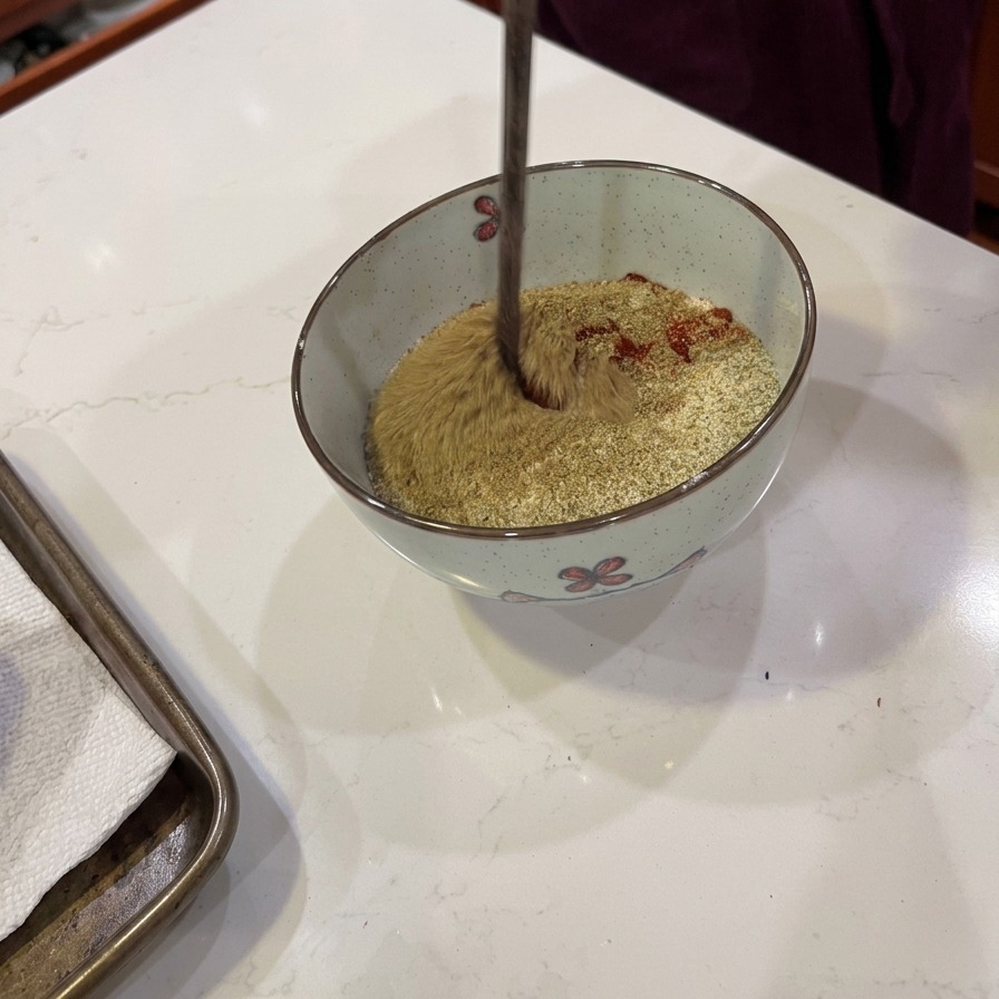A wooden utensil stirring a blend of flour and brownish-red spices in a small bowl.