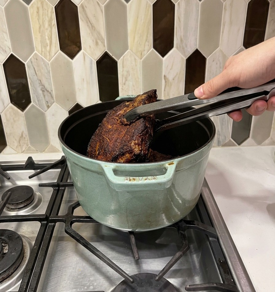 Heavily seared beef chunks being transferred with metal tongs into a light green cast iron Dutch oven.