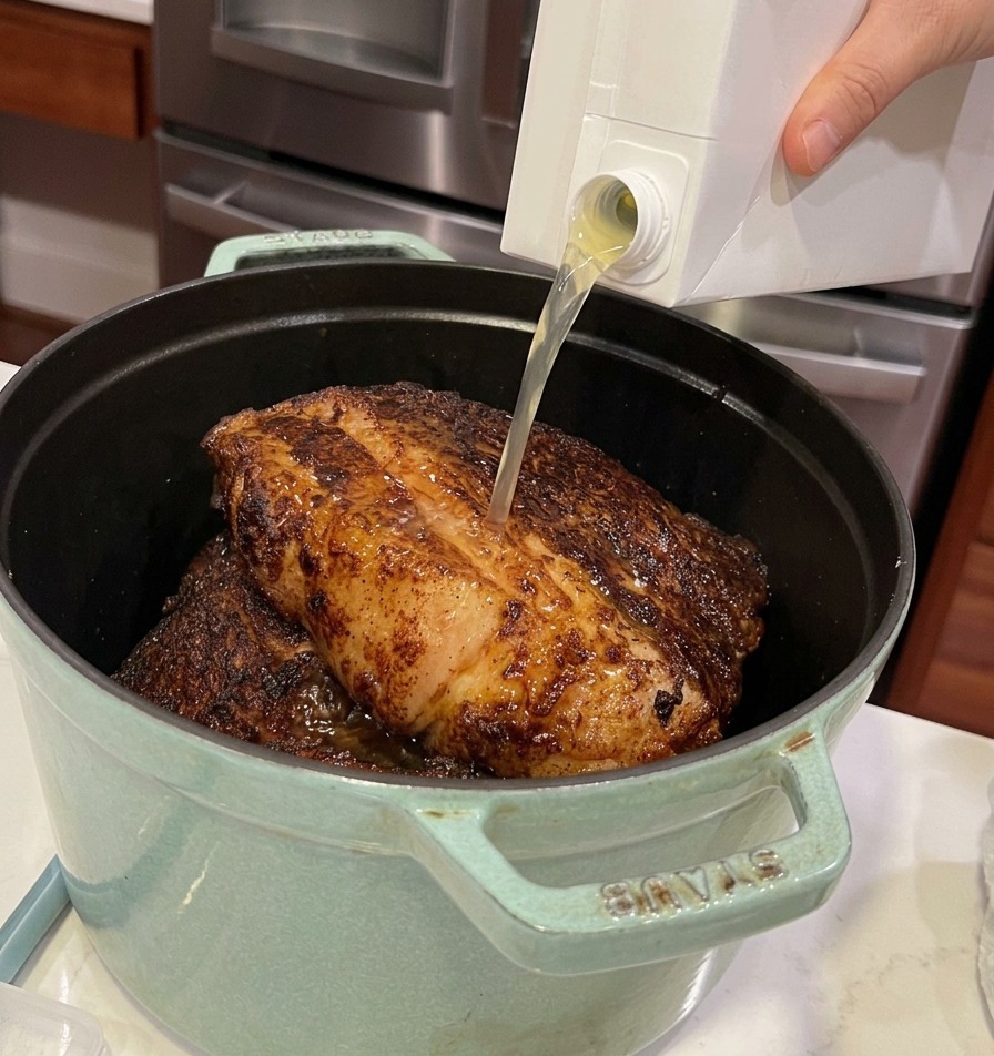 Pouring light-colored chicken stock from a carton into a green cast iron pot filled with large seared beef chunks.