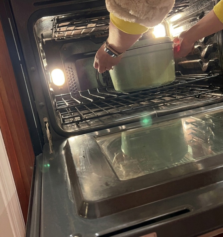 A person carefully placing a covered green cast iron Dutch oven onto the center rack of a preheated oven.