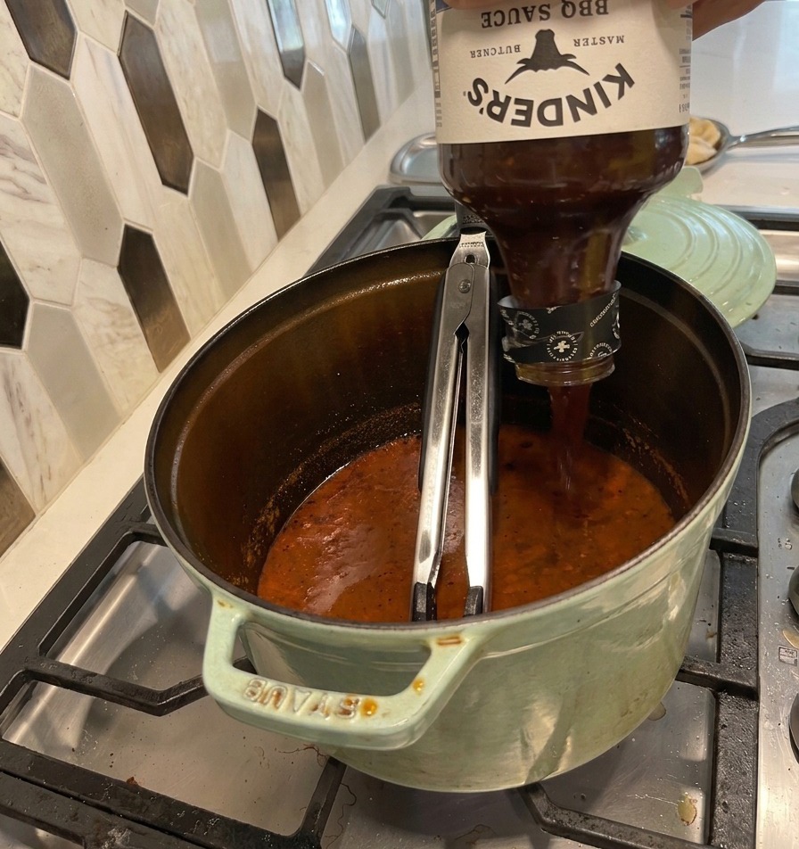 Pouring a large bottle of BBQ sauce into a green pot filled with a dark, reduced, simmering savory liquid.