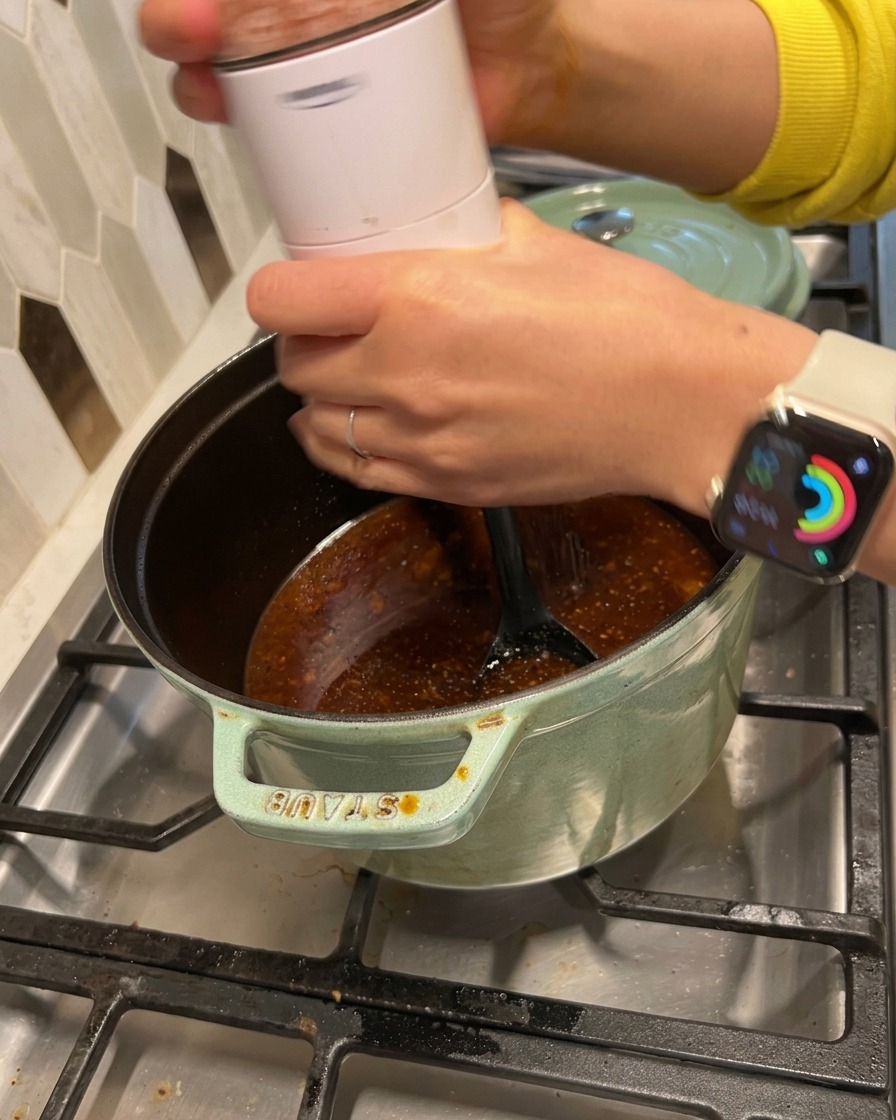 A person grinding salt and black pepper from a white electric mill into a simmering dark sauce in a green cast iron pot.