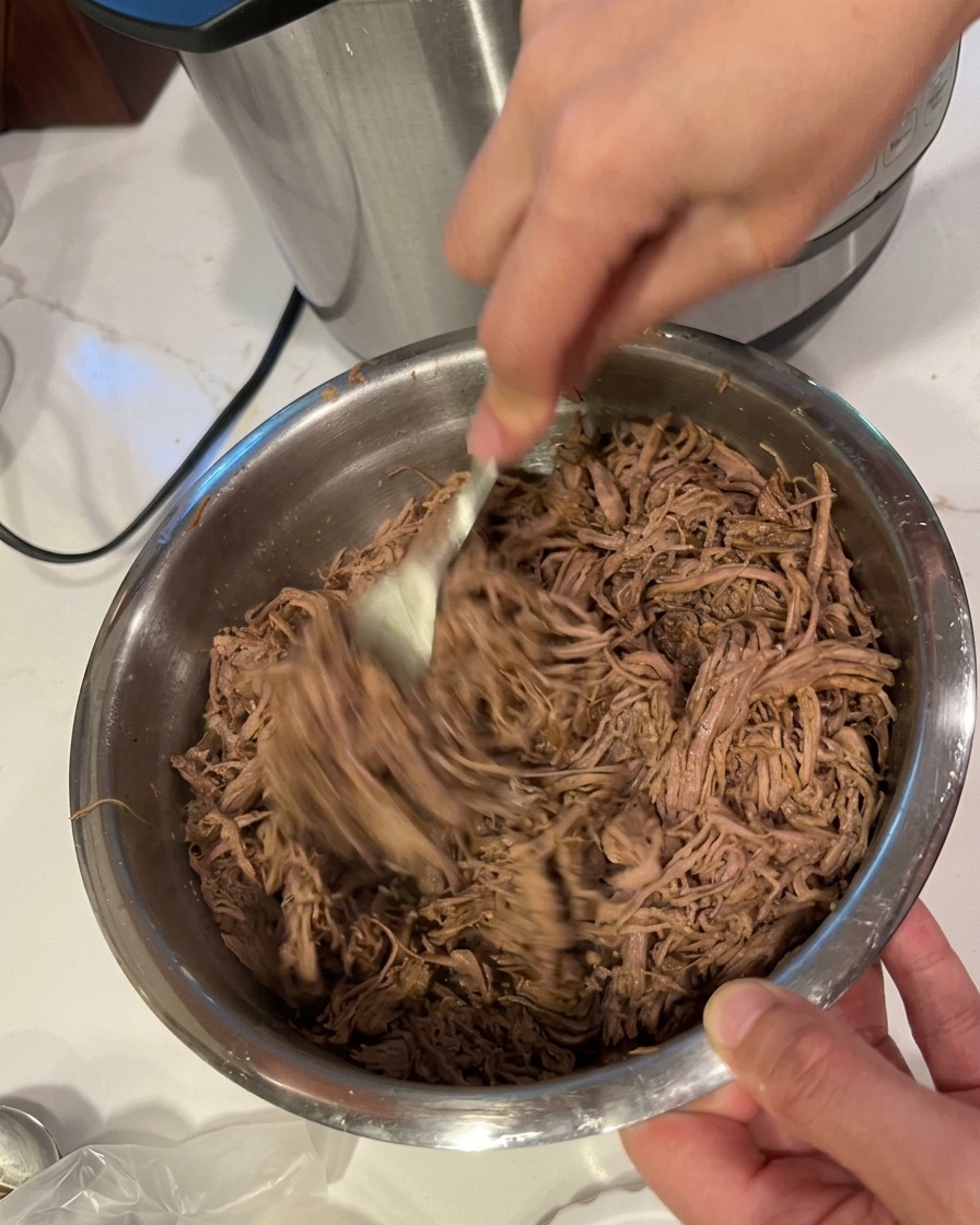 A pair of hands using a fork to shred tender, cooked beef into fine strands inside a large stainless steel bowl.