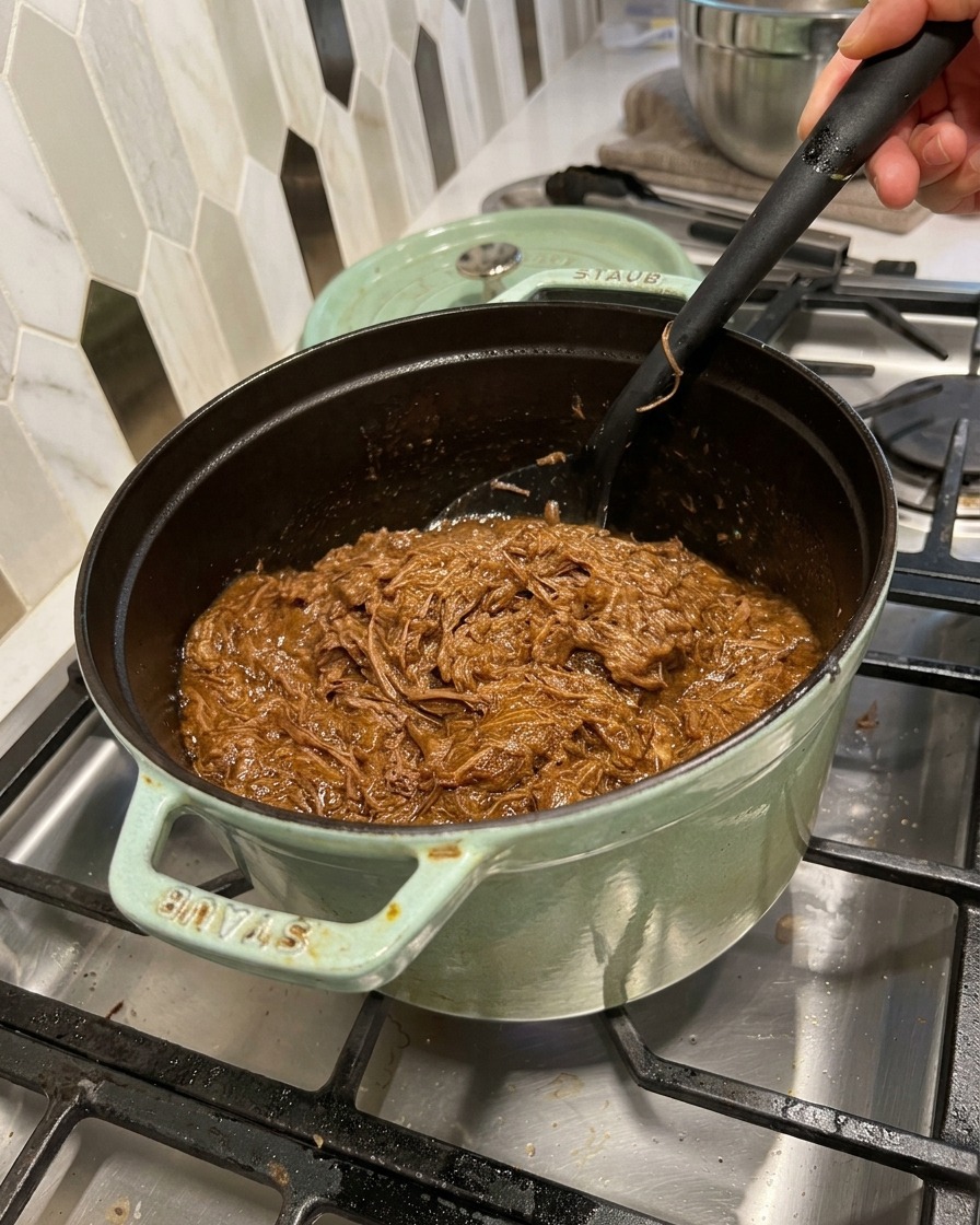 Shredded beef being stirred into a rich, dark barbecue sauce inside a green cast iron pot with a black spatula.