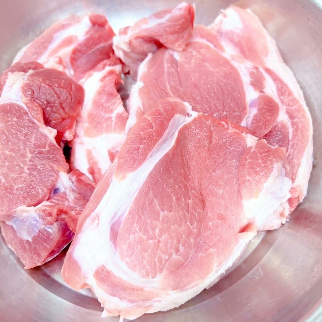 Thick cuts of raw pork shoulder placed inside a stainless steel mixing bowl.