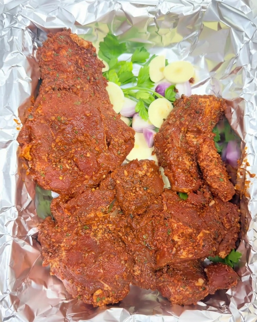 Marinated red pork chunks placed on a sheet of aluminum foil over fresh parsley, sliced shallots, and garlic.