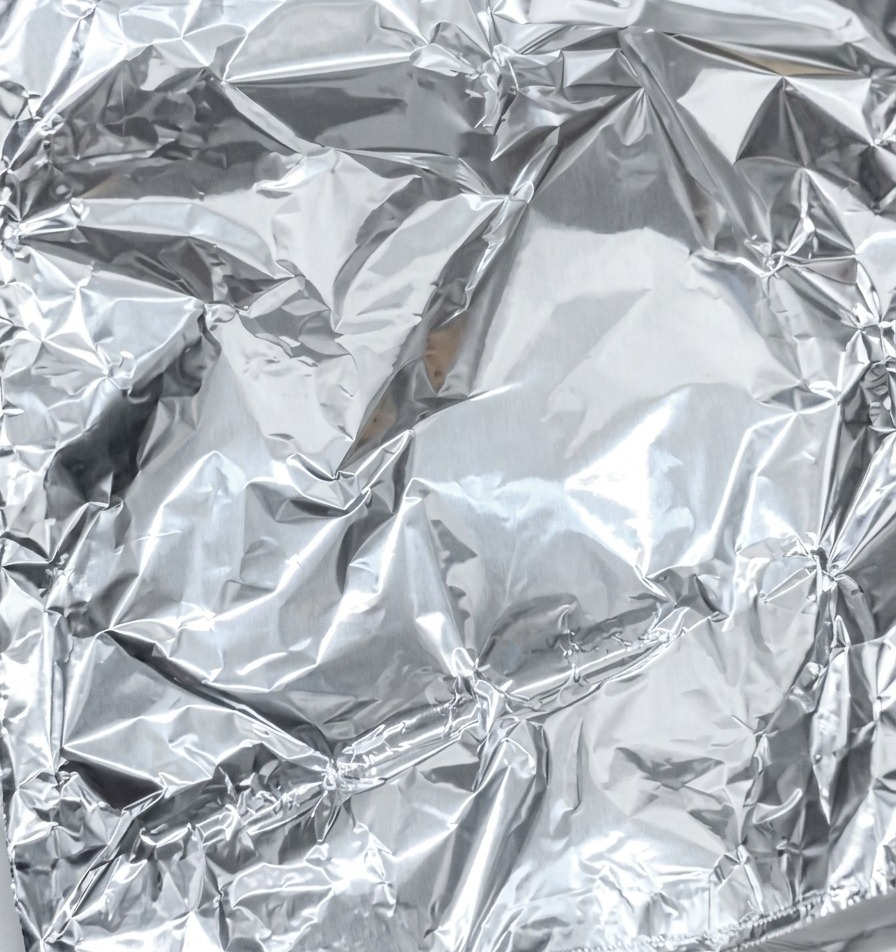 A close-up shot of shiny aluminum foil being folded and sealed to wrap a piece of marinated pork.