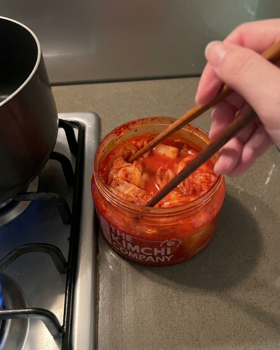 Wooden chopsticks lifting a clump of red, spicy kimchi out of a clear plastic jar.