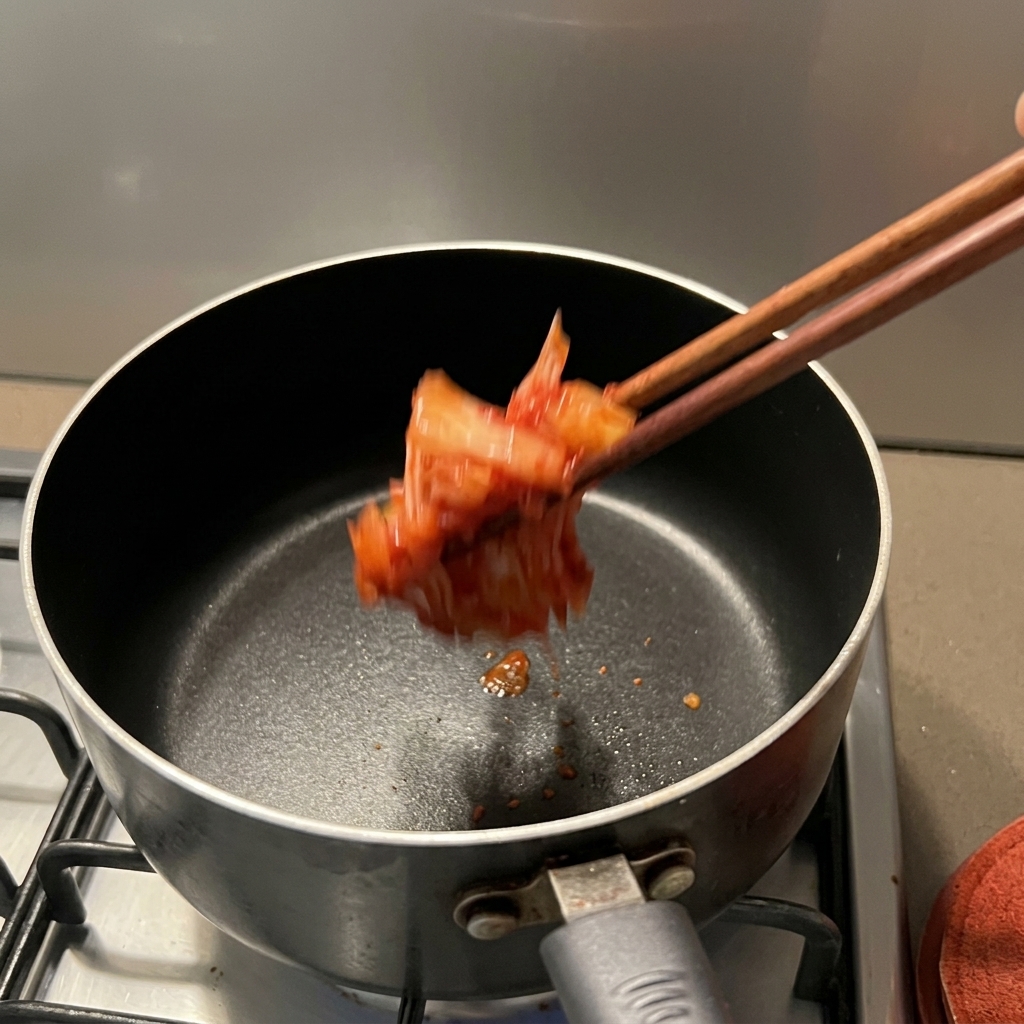 A clump of bright red, seasoned kimchi being dropped into a dark non-stick saucepan using wooden chopsticks over a stovetop.