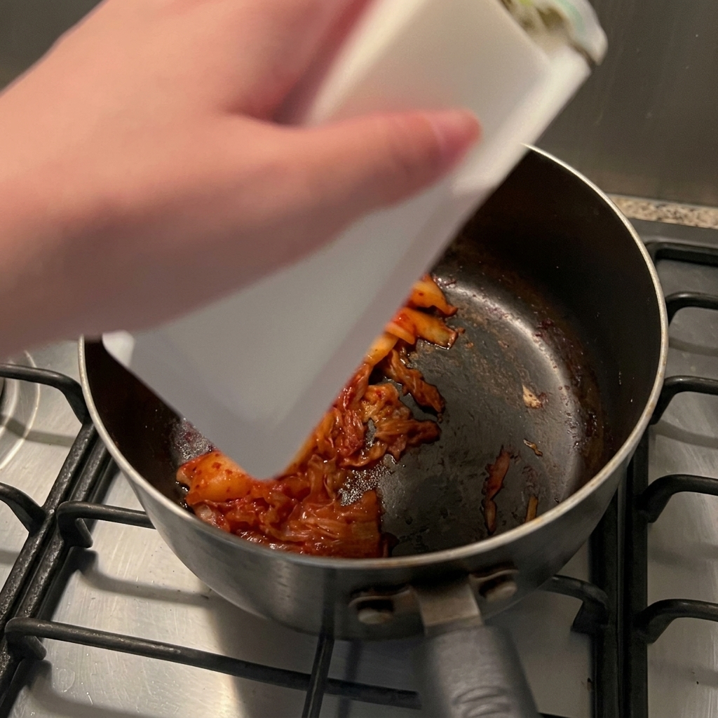 A block of white silken tofu being slid from its plastic container into a dark saucepan containing sautéed kimchi.