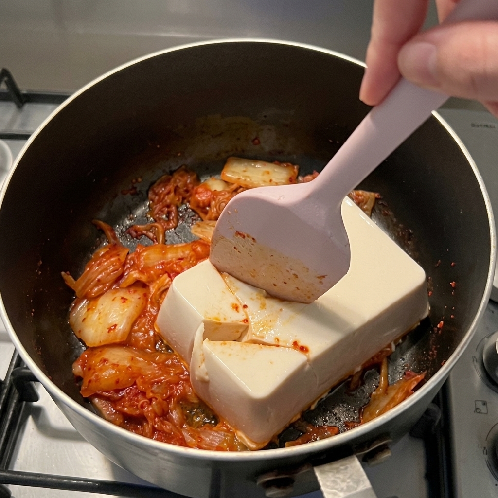 A pink silicone spatula slicing down through a block of white silken tofu over red kimchi in a dark saucepan.