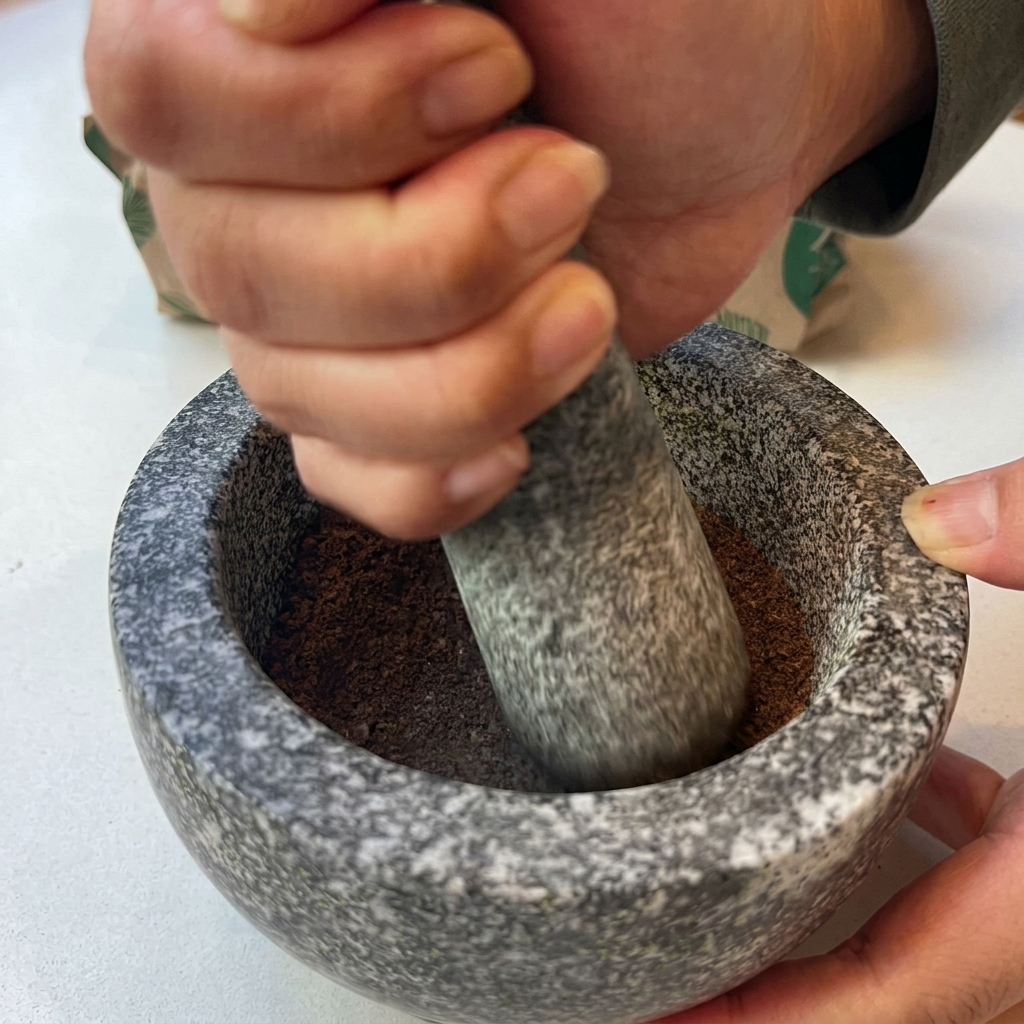 A pair of hands using a stone pestle to grind toasted spices into a fine brown powder inside a granite mortar.
