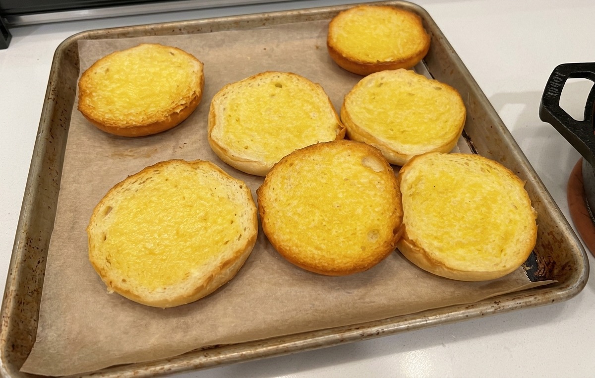 Buttered halves of hamburger buns laid out on parchment paper on a baking tray.