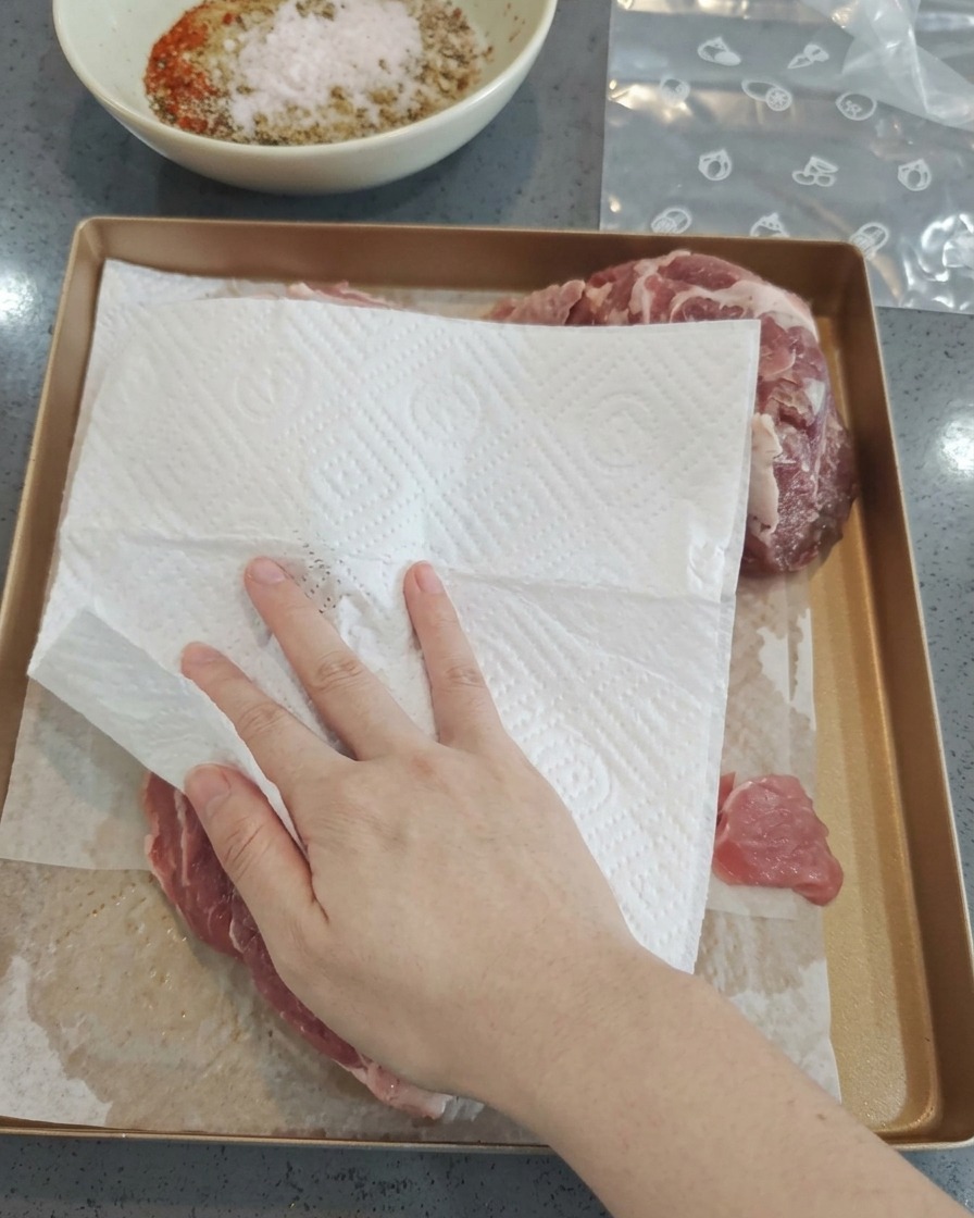 A hand using a white paper towel to pat dry raw pork pieces on a gold-colored baking tray.