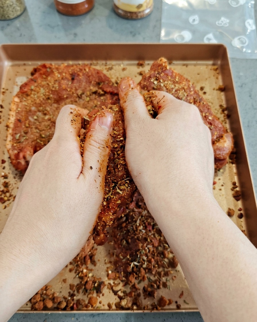 Hands firmly rubbing a dark, savory spice blend onto large pieces of raw pork on a baking tray.
