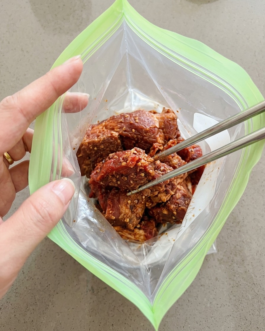 Seasoned pork chunks being placed into a green reusable zip-top bag using metal chopsticks.