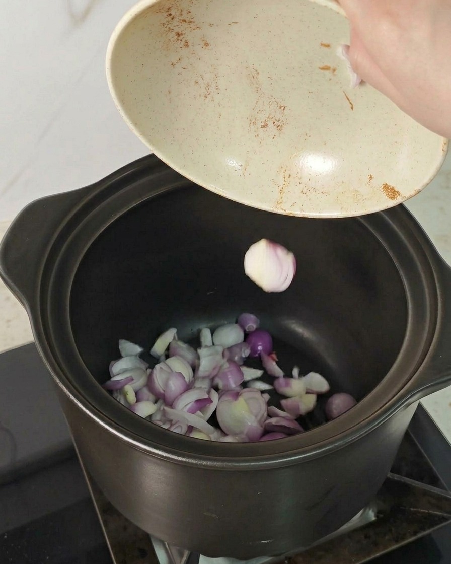 Slices of red shallots being poured from a bowl into the bottom of a dark ceramic cooking pot.