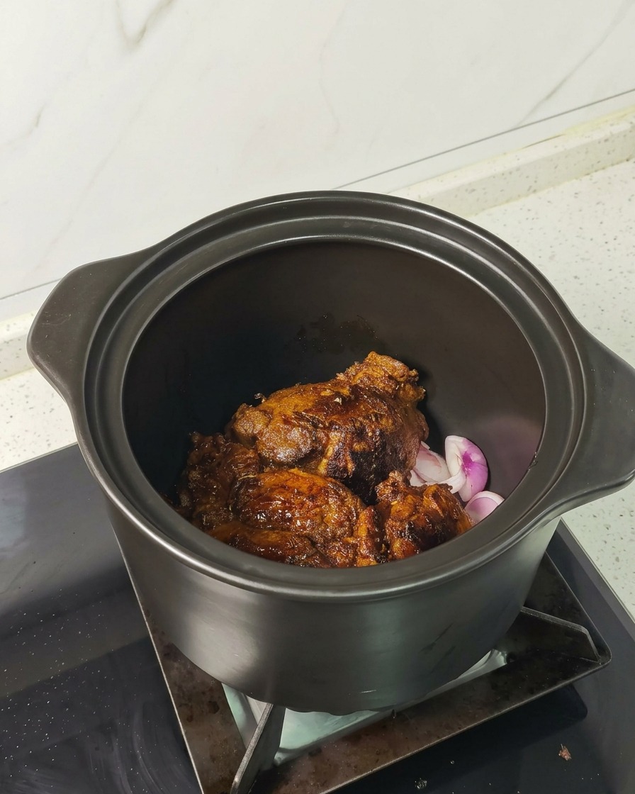 Deeply browned, seared pork chunks resting on sliced red onions inside a dark ceramic braising pot.
