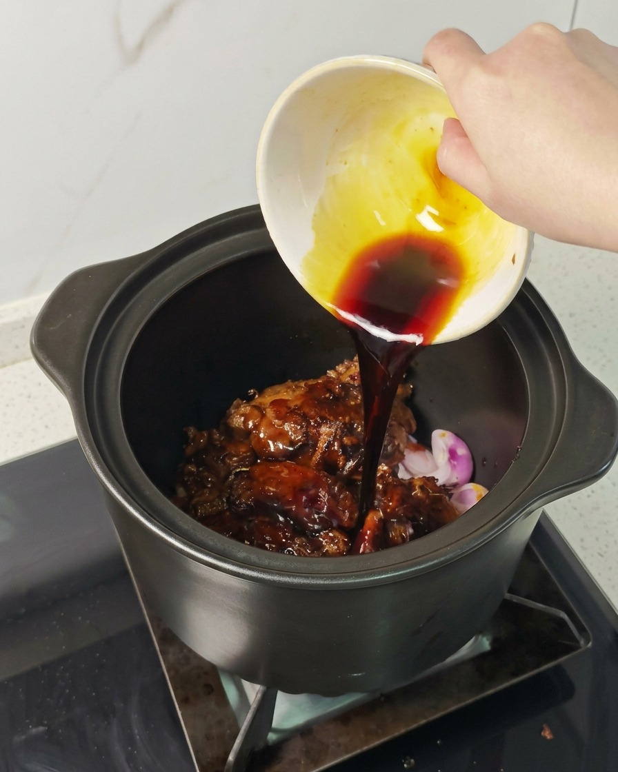 A hand pouring a dark, savory sauce from a small bowl over seared pork in a cooking pot.