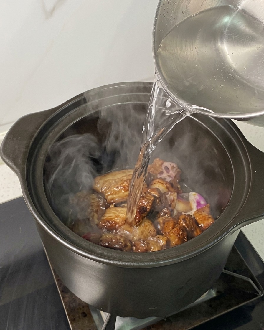 Steaming hot water being poured from a kettle over braising pork and onions in a pot.