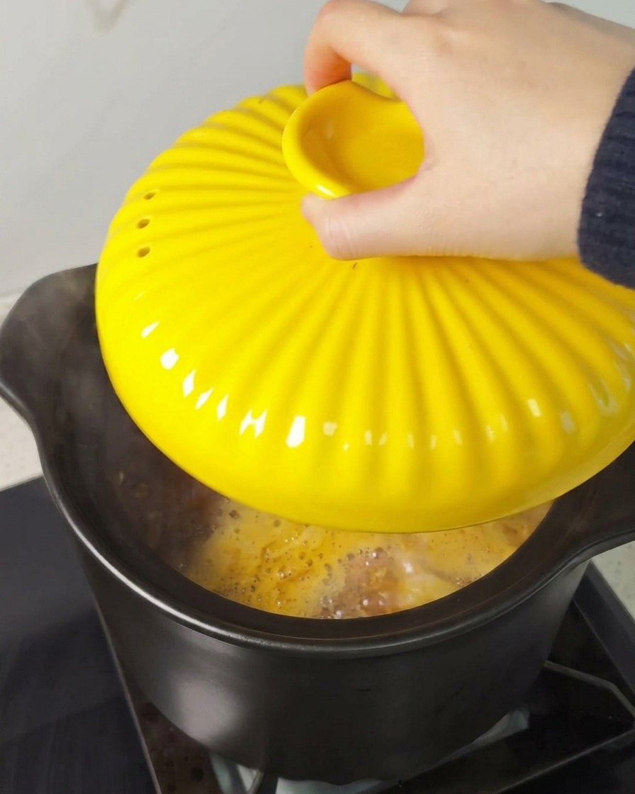 A hand placing a bright yellow, fluted lid onto a black braising pot over a stovetop.
