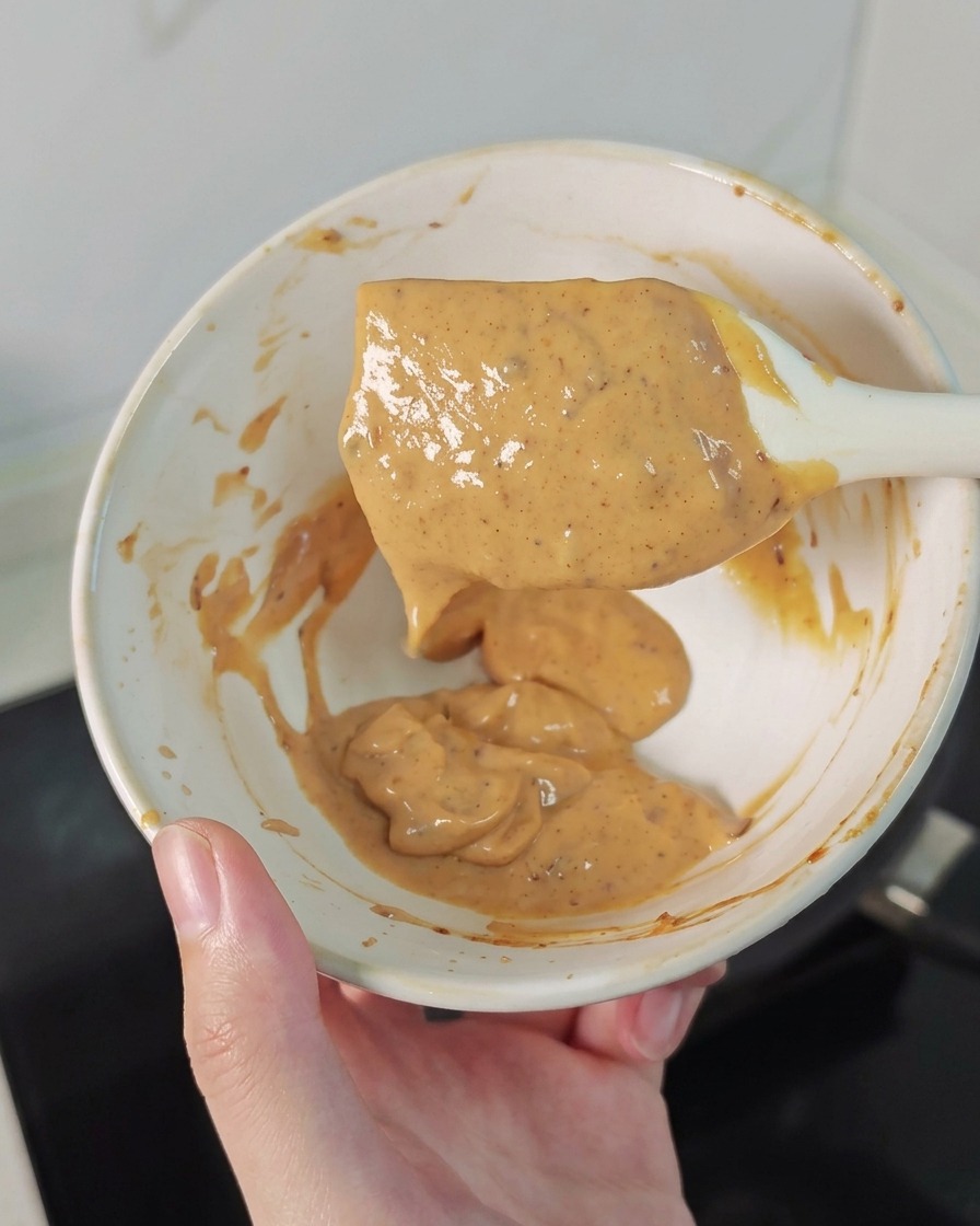 A hand holding a small bowl and a white spatula coated in a creamy, speckled orange sauce.