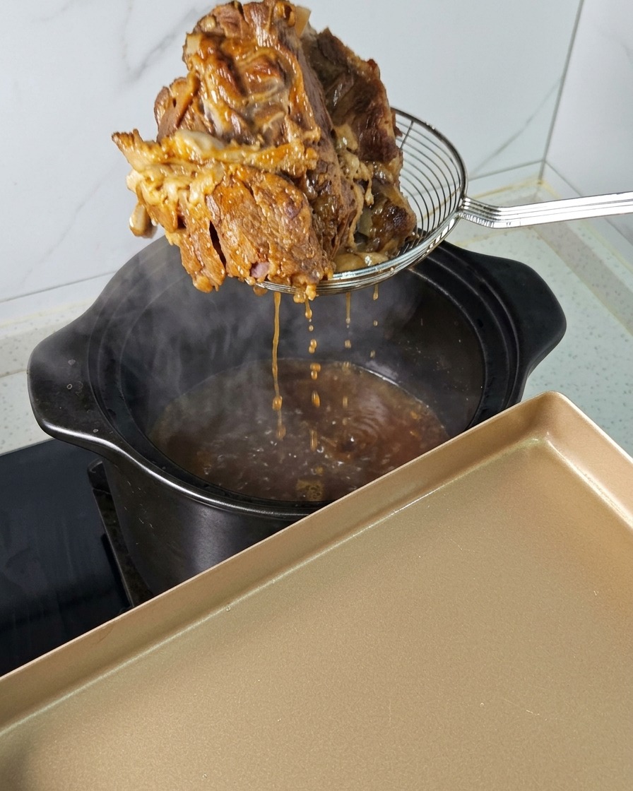 A large piece of tender, braised pork being lifted out of a steaming black pot with a wire spider strainer.
