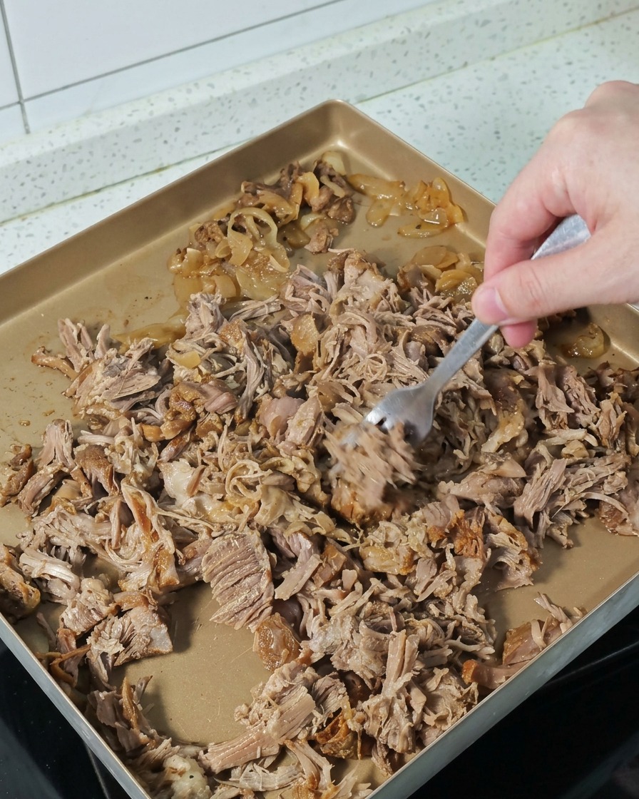 A hand using a fork to pull apart and shred pieces of tender, cooked pork on a gold-colored baking tray.