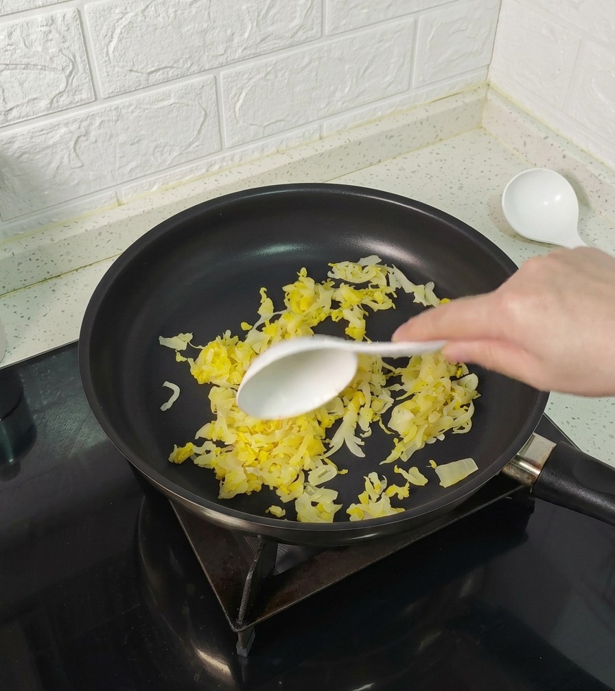 A hand using a white spoon to stir shredded yellow sauerkraut in a black non-stick frying pan.