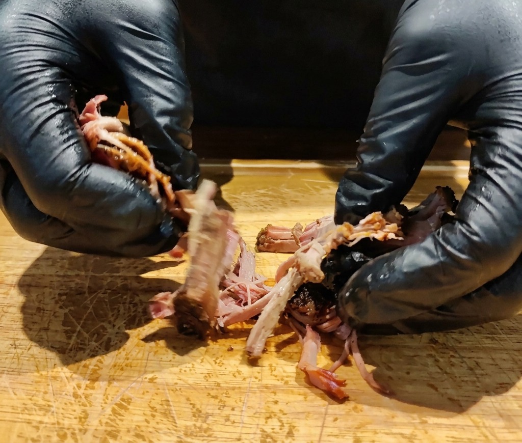 Gloved hands pulling apart a dark, slow-roasted piece of pork collar on a wooden cutting board.