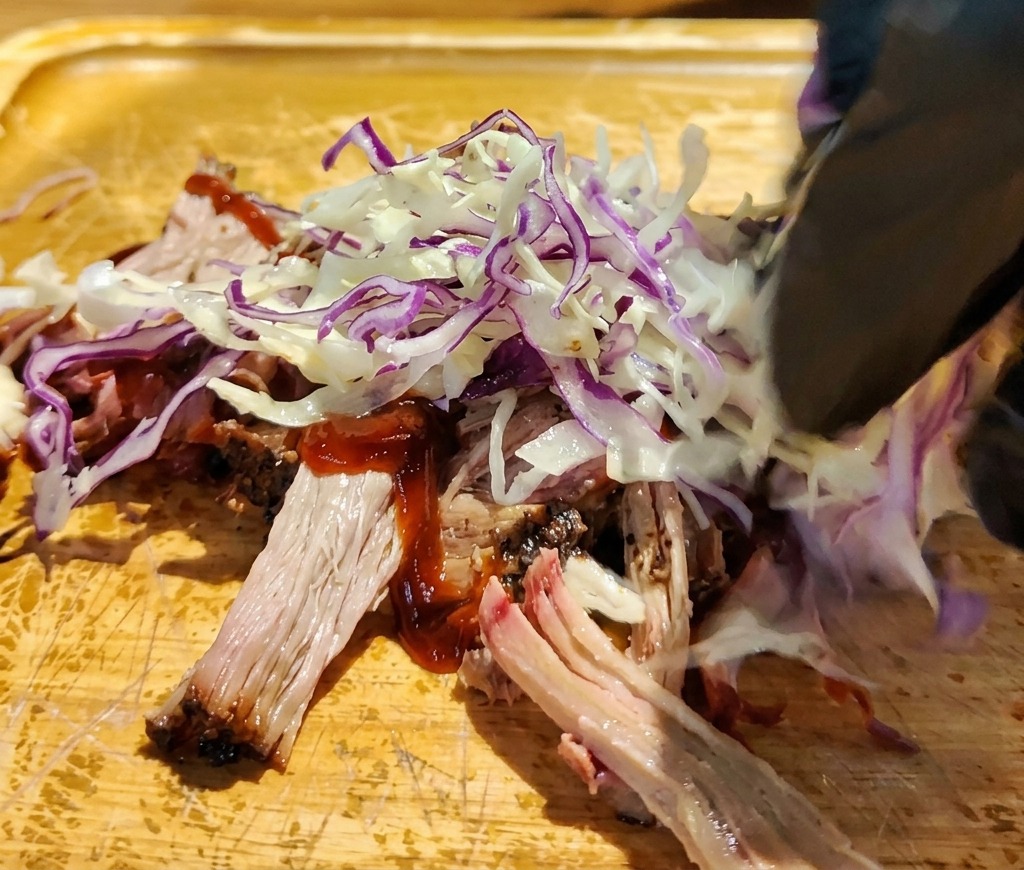 Gloved hand placing shredded red and white cabbage over sauced pulled pork on a wooden cutting board.