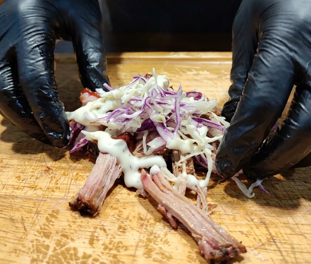 Gloved hands beside a pile of pulled pork topped with coleslaw and creamy white dressing on a cutting board.