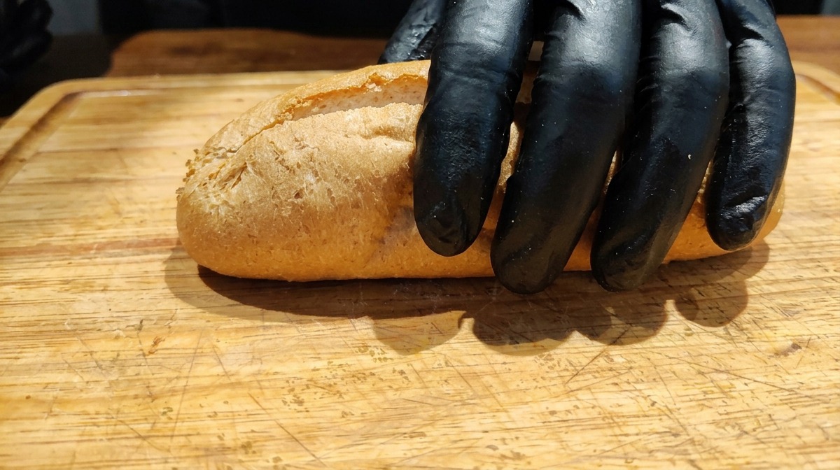 Gloved hand placing a partially sliced whole wheat baguette on a wooden cutting board.