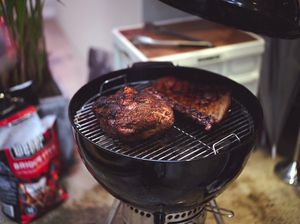 Partially cooked pork shoulder and ribs with a dark caramelized crust sitting on a grill grate.