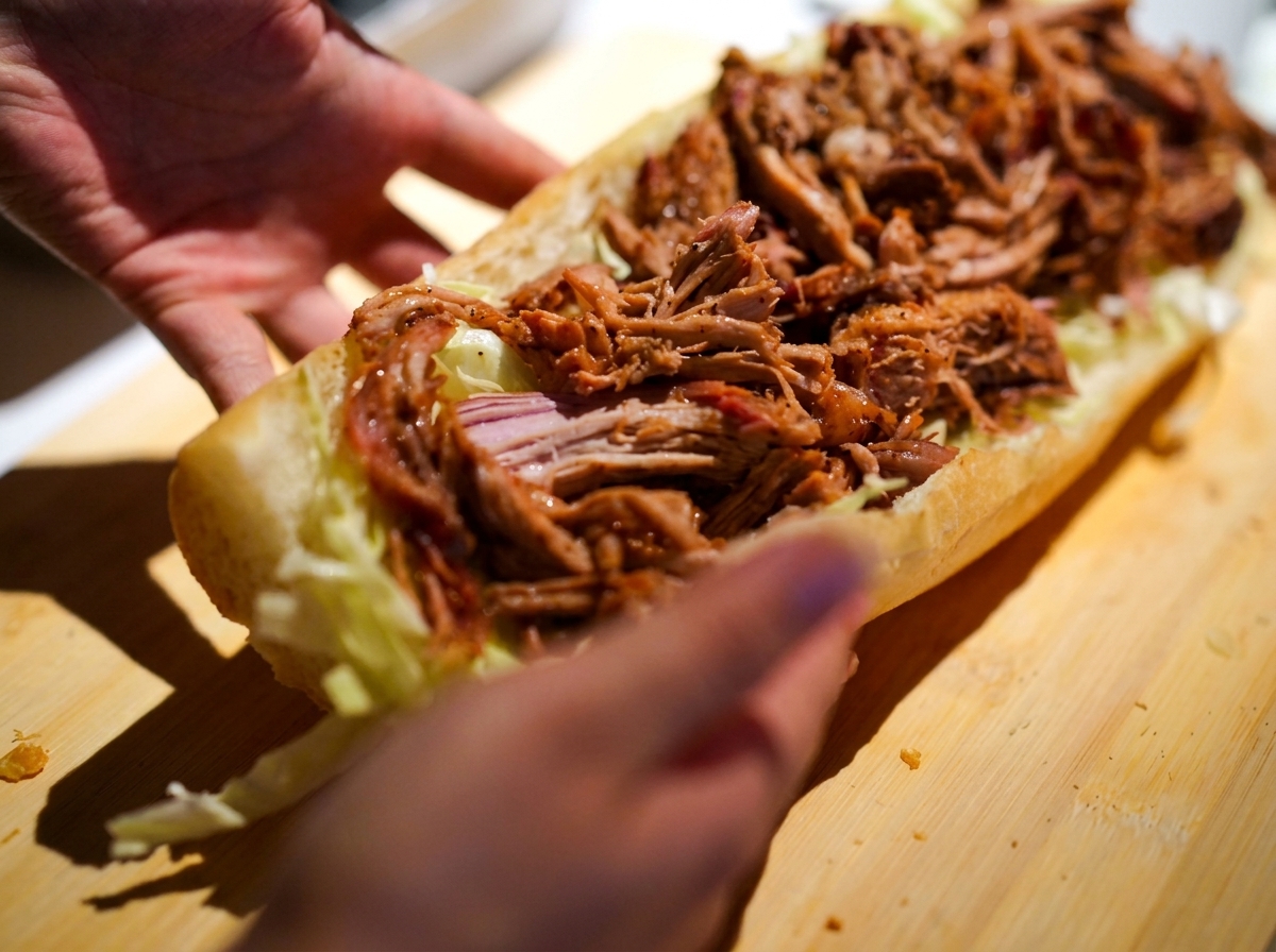 Hands carefully holding a long sandwich roll that is generously filled with creamy coleslaw and warm, saucy pulled pork.