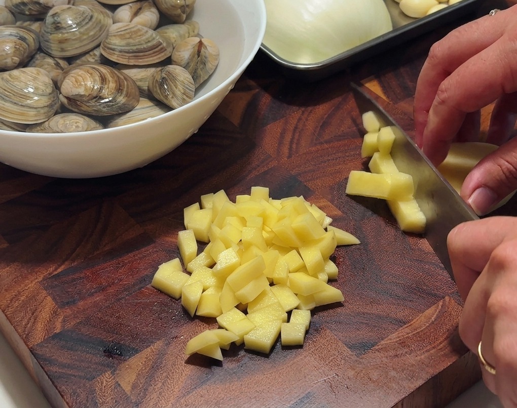 A pair of hands using a sharp chefs knife to dice peeled potatoes into small cubes on a dark wooden cutting board.