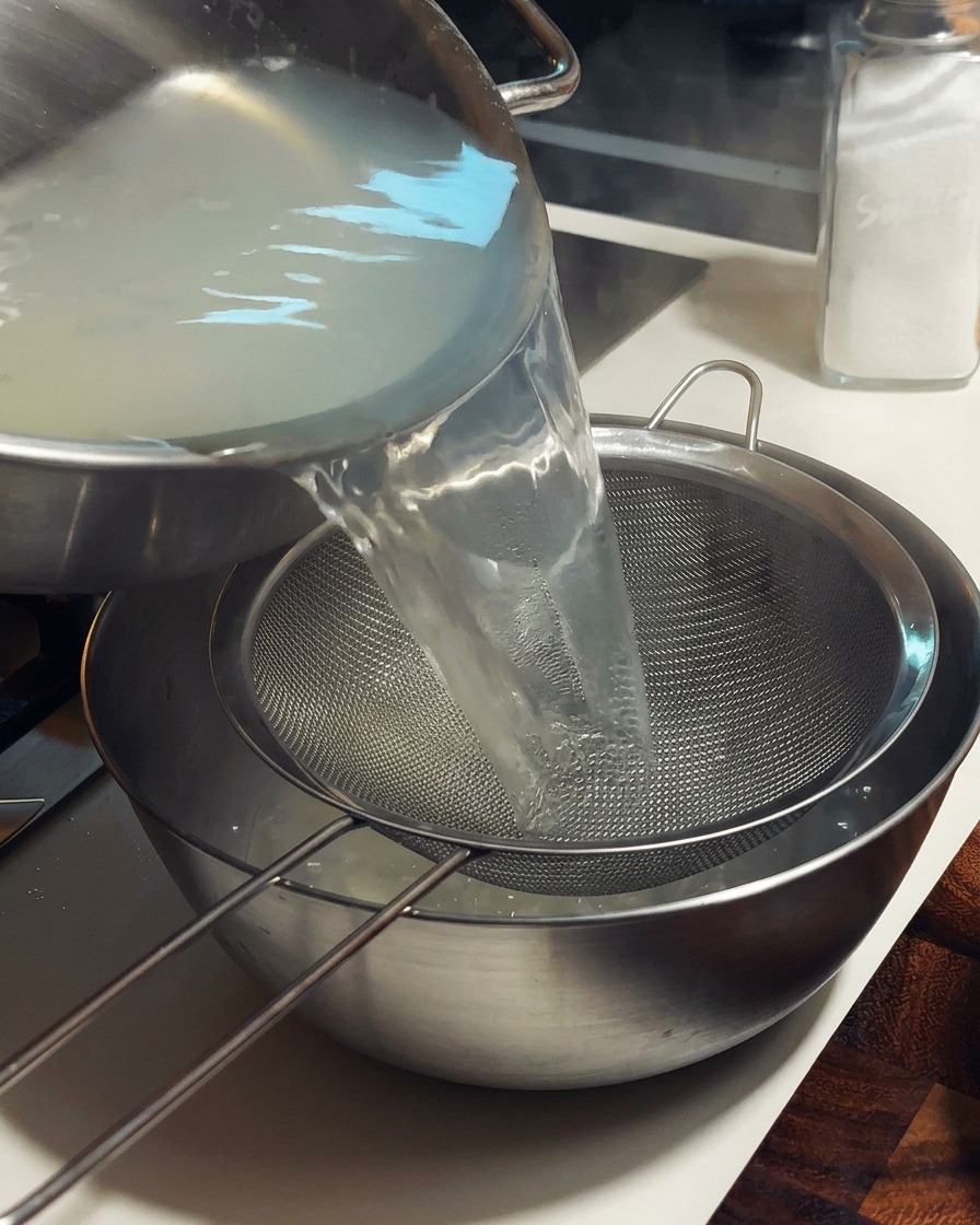 Hot, cloudy clam broth being poured from a stainless steel pot through a fine wire mesh strainer into a large metal bowl.