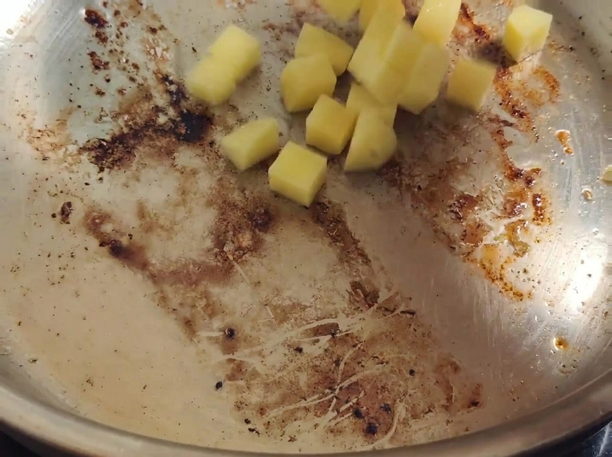 Diced yellow potatoes falling into a hot stainless steel pan coated with rendered bacon fat and dark browned bits.