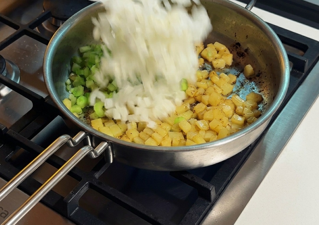 Diced white onions being added from a white plate into a stainless steel skillet containing diced celery and golden brown sautéed potatoes.