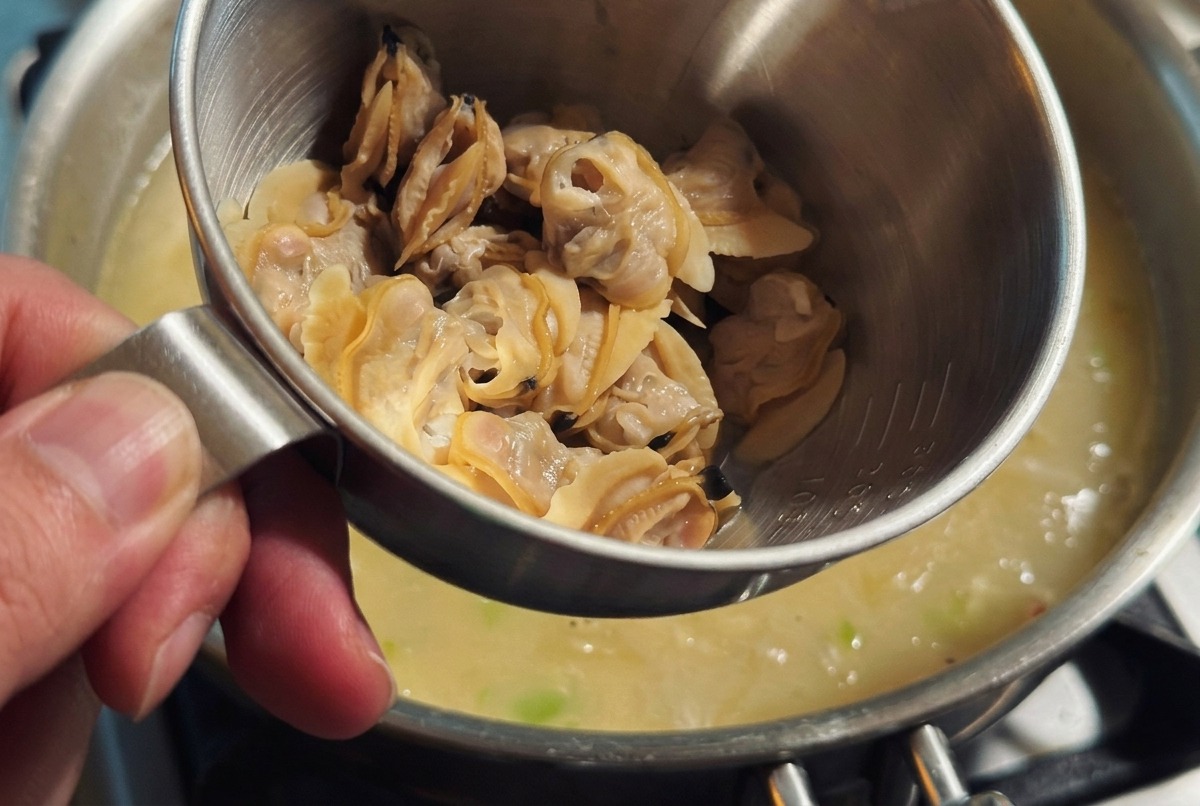 Heavy cream being poured into a pot of thick chowder while fresh clam meat is gently stirred in.