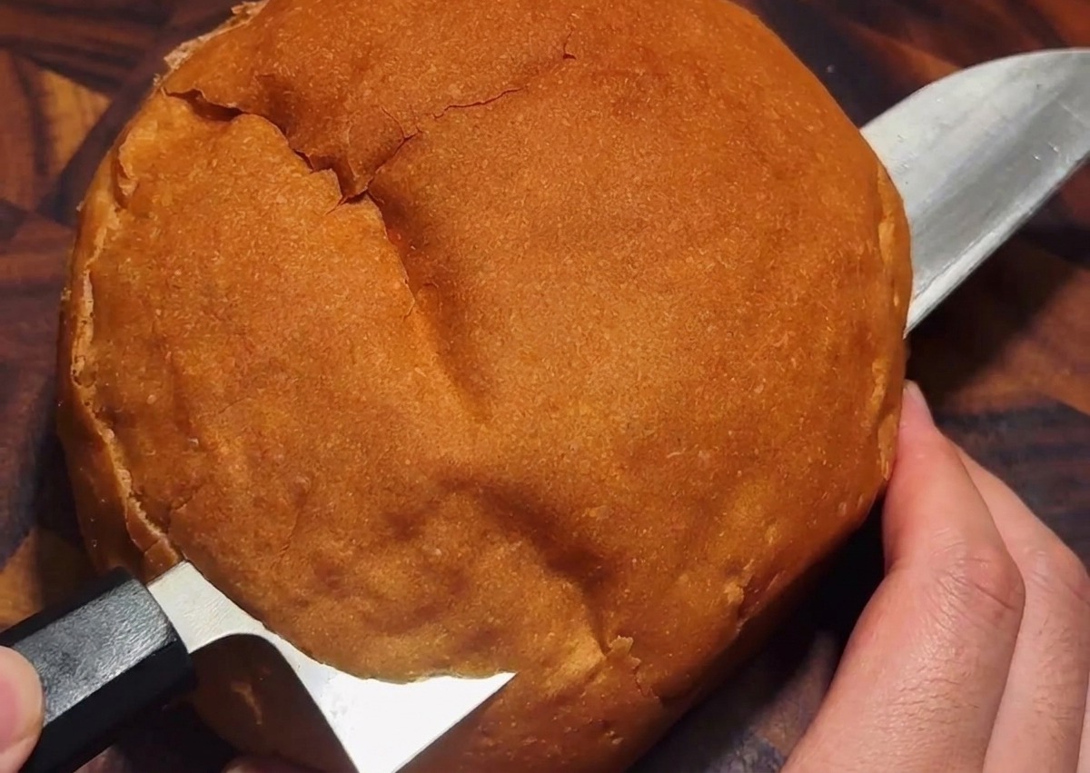 A knife cutting a circular lid into the crusty top of a large round bread loaf on a wooden surface.