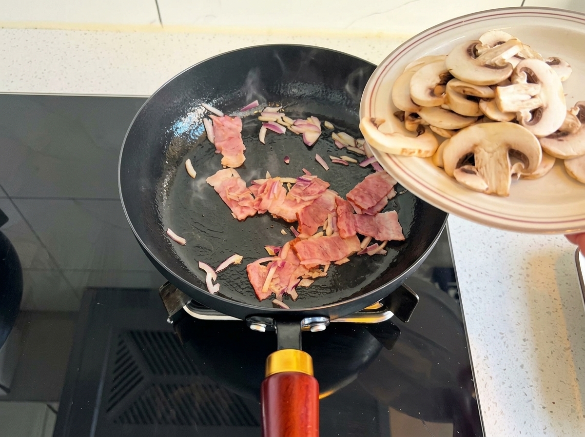Sliced white button mushrooms being slid off a plate into a frying pan with cooked onions and bacon.