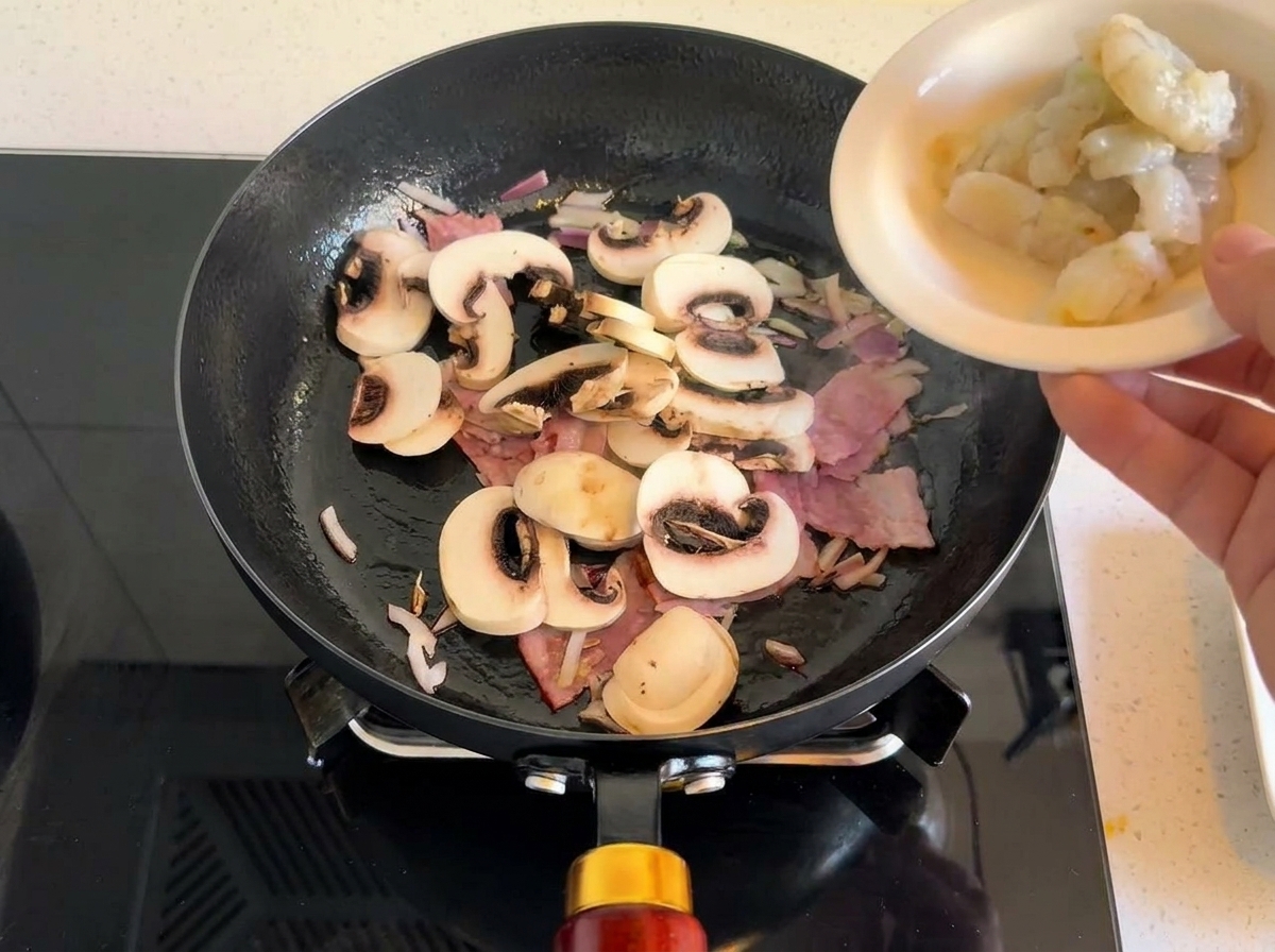 Raw grey shrimp being added to a pan with sautéed bacon, mushrooms, and onions.