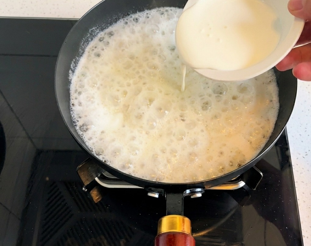 Thick white heavy cream being poured from a small bowl into a simmering pan of white soup base.
