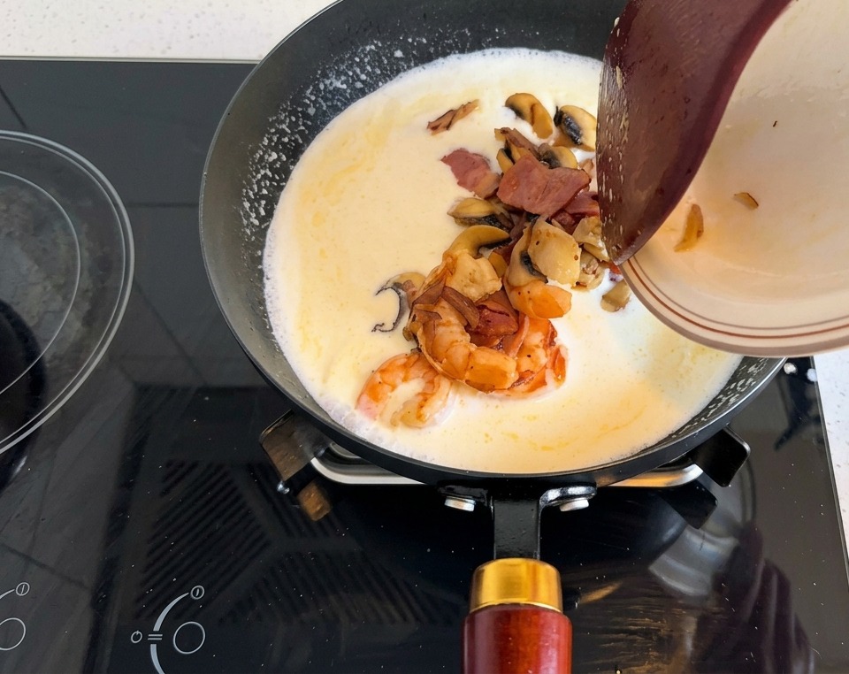 A plate of cooked shrimp, bacon strips, and sliced mushrooms being tilted into a creamy soup base in a black pan.