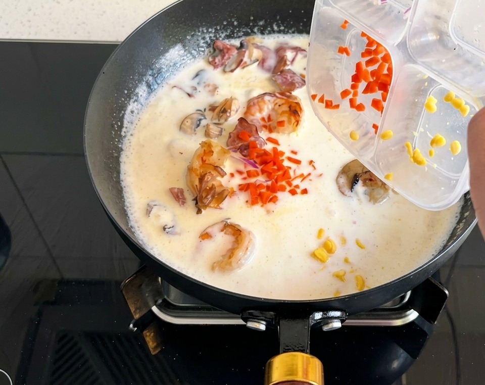 Diced orange carrots and yellow corn kernels being poured from a plastic container into a pan of bubbling cream soup.