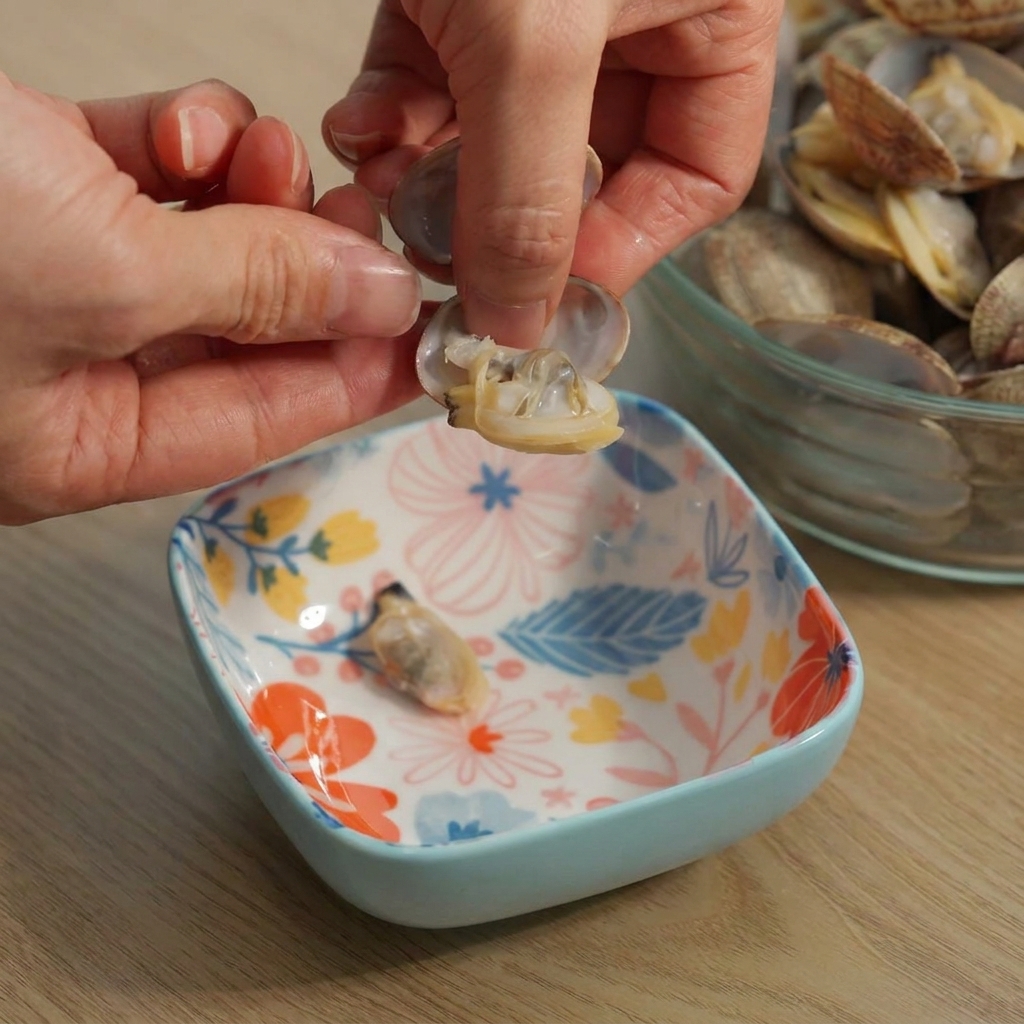 Hands carefully pulling cooked clam meat out of an open shell over a small patterned bowl.