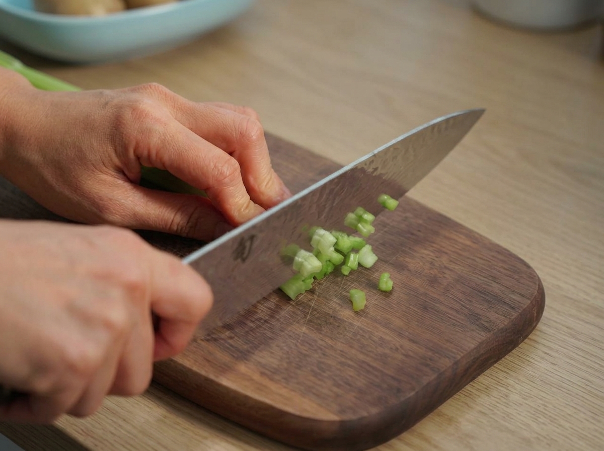 Hands holding a large chefs knife, dicing bright green celery stalks on a wooden cutting board.